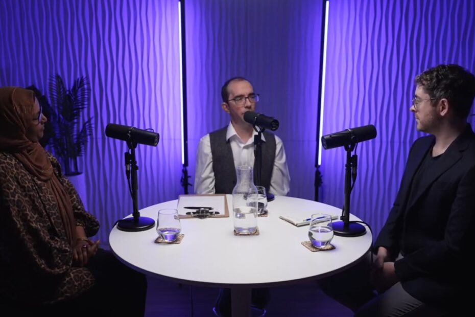 Three speakers sit spotlighted around a round white table, with a purple background.
