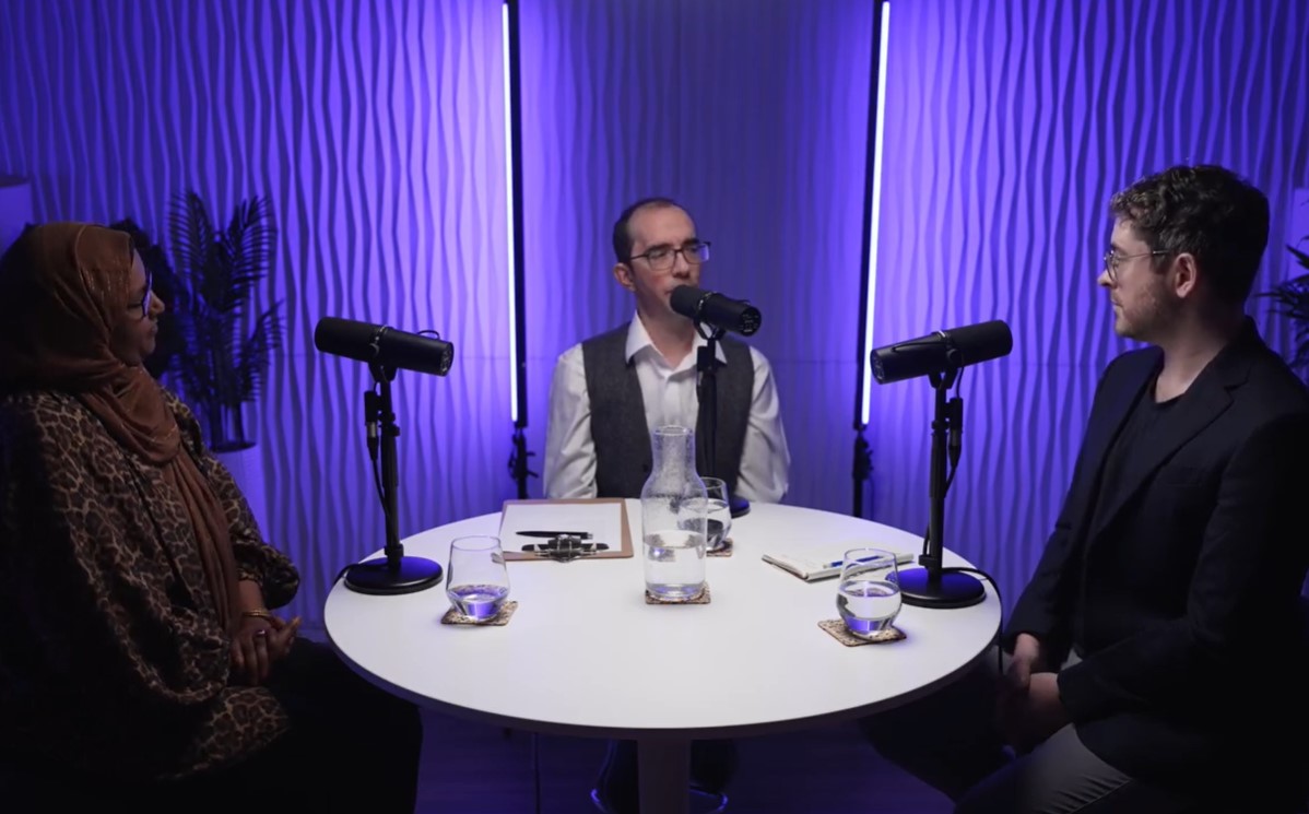 Three speakers sit spotlighted around a round white table, with a purple background.