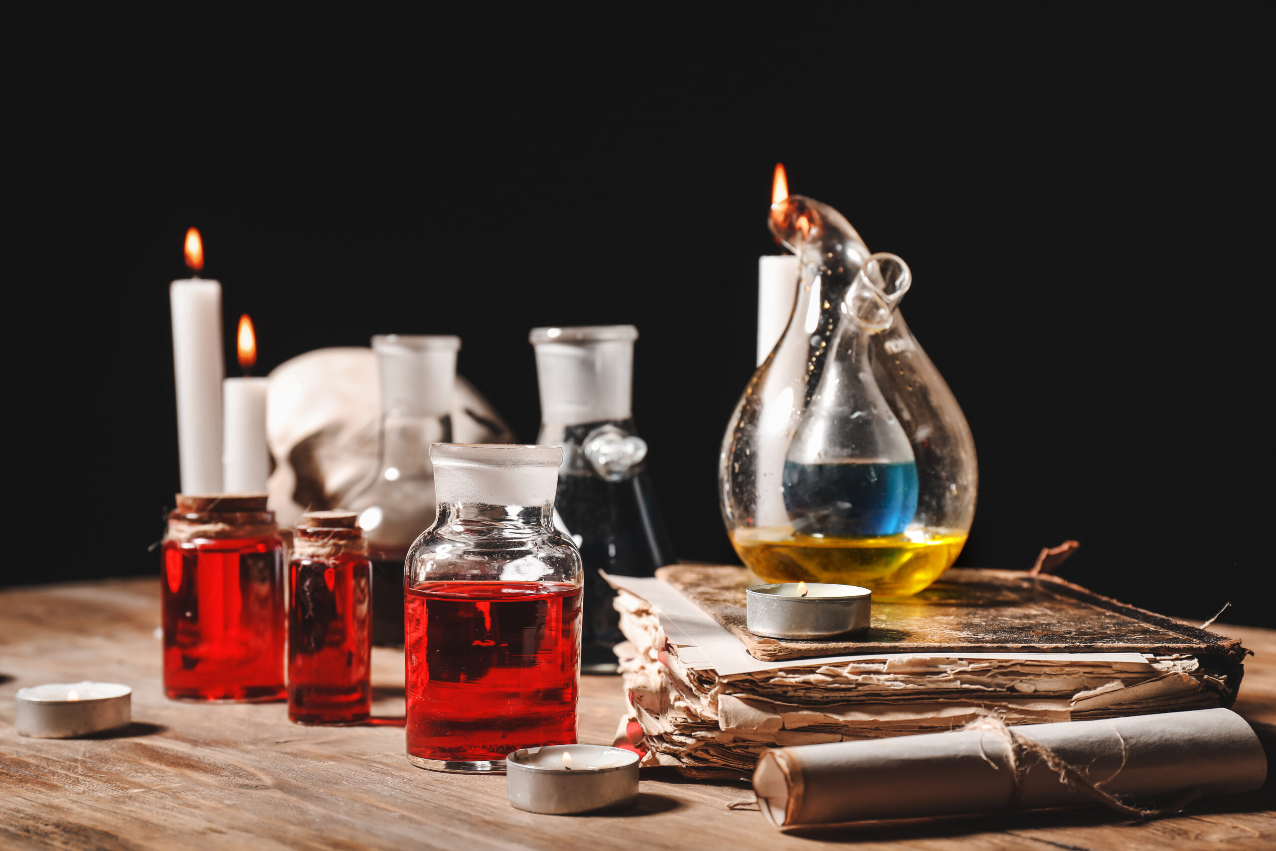 Old fashioned glass bottles containing coloured liquids are grouped next to a leather-bound book and a lit candle.